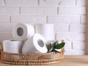 Basket with white toilet paper rolls and a few green leaves on a wooden surface against a white brick wall.